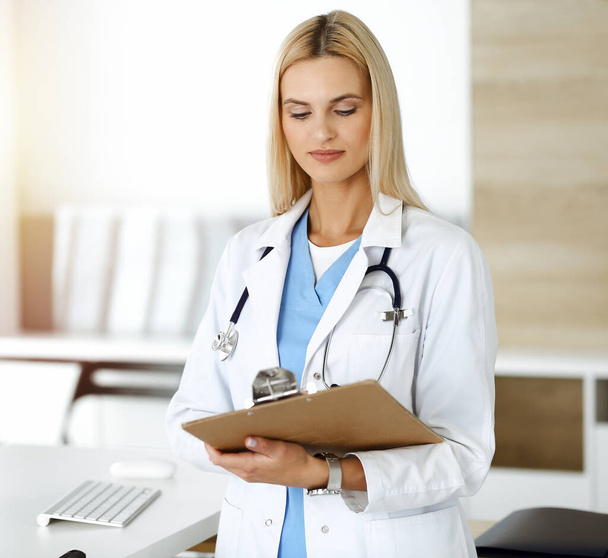 Doctor woman controls medication history records and exam results while using clipboard in sunny hospital . Medicine and healthcare concept - Foto, imagen