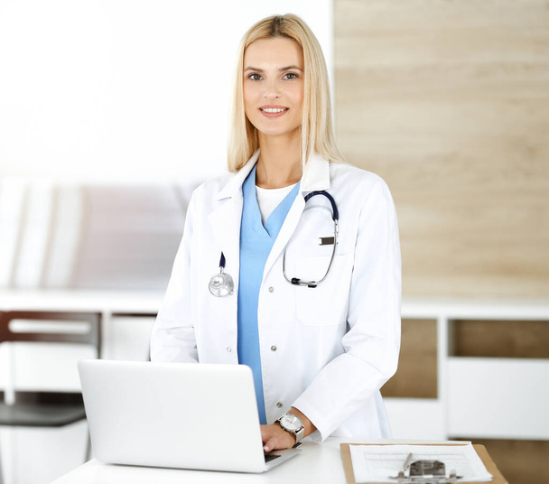 Woman-doctor controls medication history records and exam results while using laptop computer in sunny hospital. Medicine and healthcare concept - Foto, imagen