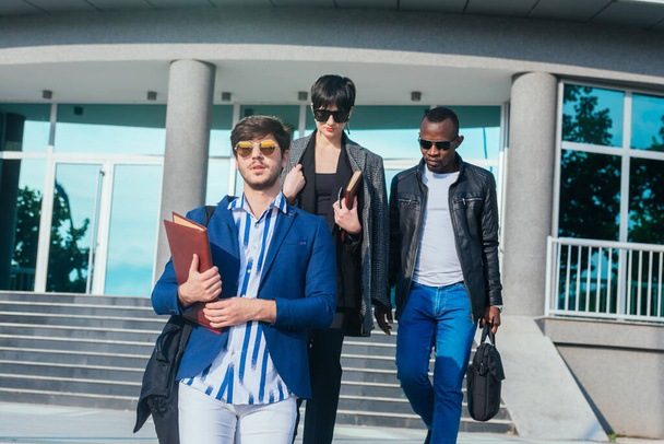 Jovens pessoas multi-étnicas modernas em desgaste formal discutindo negócios enquanto caminham fora do prédio de escritórios - Foto, Imagem