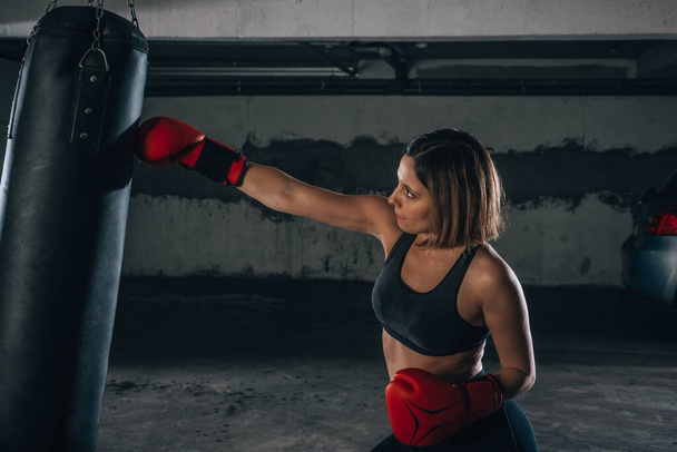 Profile view of a strong young woman punching a boxing bag inside the garage - Photo, Image