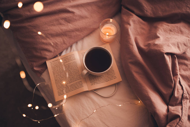 Cup of fresh hot coffee staying on open paper book with burning candle and glowing Christmas lights in bed closeup. Winter holiday season. Good morning. Selective focus. - Photo, Image
