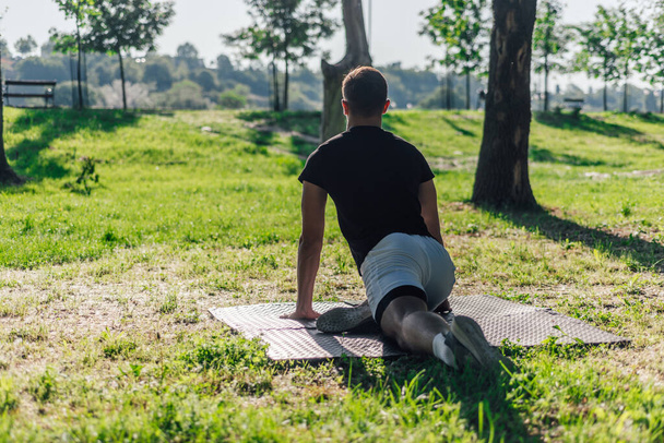 Υγιής νεαρός άνδρας κάνει εξωτερική προθέρμανση ασκήσεις τεντώματος σε ένα στρώμα γυμναστικής - Φωτογραφία, εικόνα