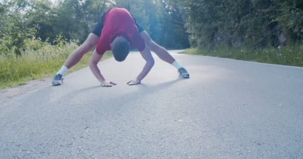 Pasujący biały mężczyzna rozciągający nogi podczas rozgrzewania się do biegania po górskiej drodze ze słońcem świecącym za nim - Materiał filmowy, wideo