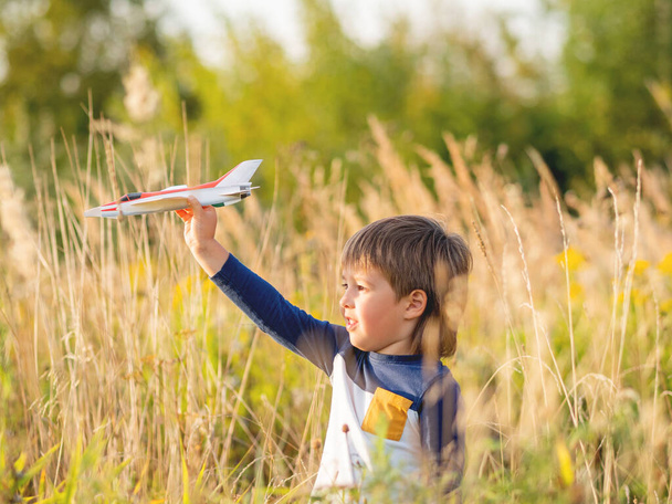 Cute toddler plays with toy airplane on field. Happy kid dreams to be a pilot. Boy is planning for the future. Golden sunset hour at autumn season. - Фото, изображение