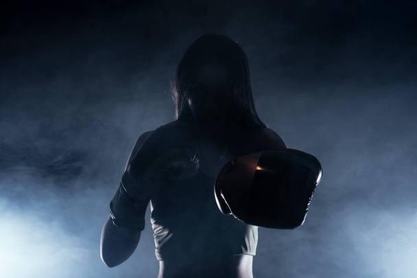 Tmavě kontrastní fotografie s kouřem v pozadí zaostřené silné ženy s boxerskými rukavicemi cvičící údery - Fotografie, Obrázek