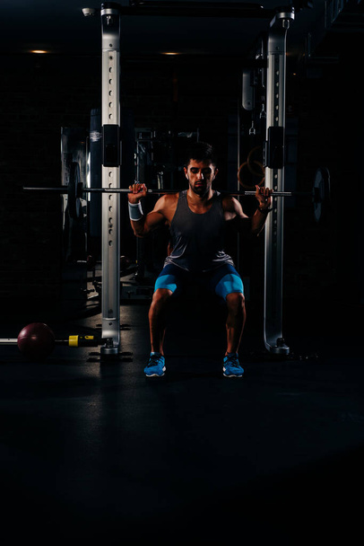 Spor salonunda ağırlık kaldıran zayıf kaslı adam, yüksek kontrastlı, koyu renk görüntü - Fotoğraf, Görsel