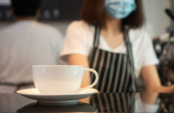 Focus selettivo della tazza di caffè e lo sfondo della donna barista asiatica che indossa maschere facciali Per prevenire malattie contagiose e la preparazione di caffè nella caffetteria. Il concetto di prevenzione del COVID 19 - Foto, immagini
