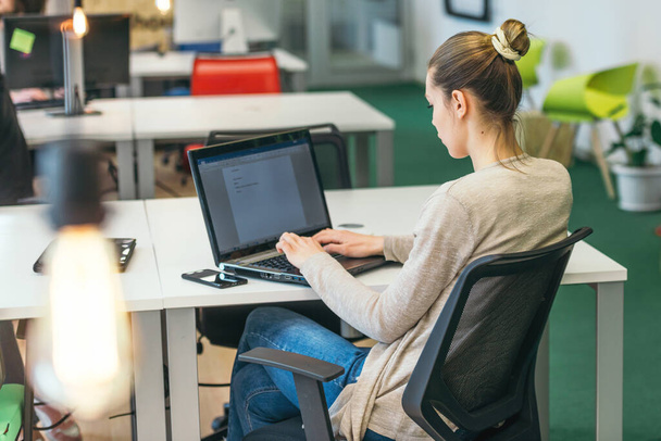 Skoncentrowana piękna dziewczyna z długimi czarnymi włosami i żółtą bluzką pracująca na laptopie w nowoczesnym biurze. - Zdjęcie, obraz