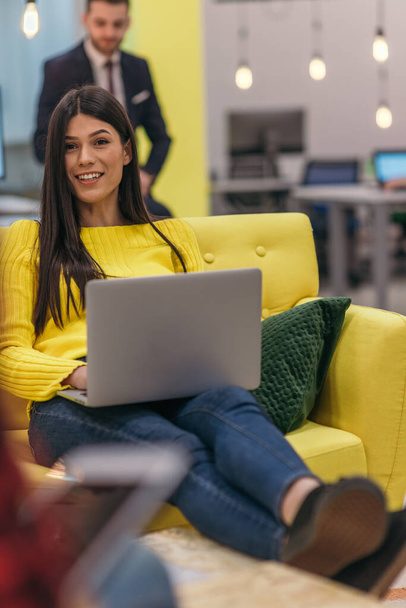 Bonita empresária (funcionária) sentada em um sofá amarelo enquanto trabalhava em seu laptop na empresa startup. - Foto, Imagem