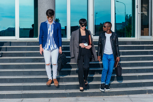 Fiatal, modern, többnemzetiségű, formális viseletű emberek üzletről beszélgetnek, miközben az irodaépületen kívül sétálnak - Fotó, kép