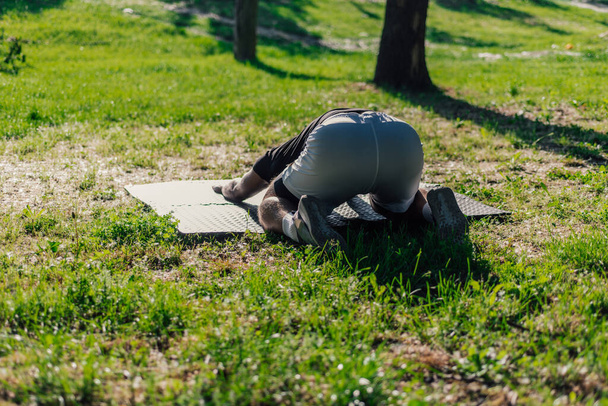 Young caucasian man practicing yoga on a mat outdoors at the park in child pose - Photo, Image