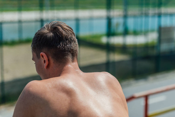 Вид сзади сильного спортсмена без рубашки, сидящего в парке - Фото, изображение
