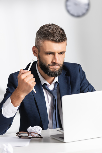 Hombre de negocios estresado sosteniendo lápiz roto cerca de gadgets, papel agrupado y anteojos en primer plano borroso en la oficina  - Foto, Imagen