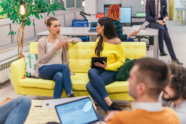 Multiethnische Kollegen blonde kaukasische und lateinamerikanische dunkelhaarige hübsche Geschäftsfrauen (Angestellte) sitzen auf einer gelben Couch, während sie an einem Laptop in einem modernen Start-up-Unternehmen arbeiten - Foto, Bild