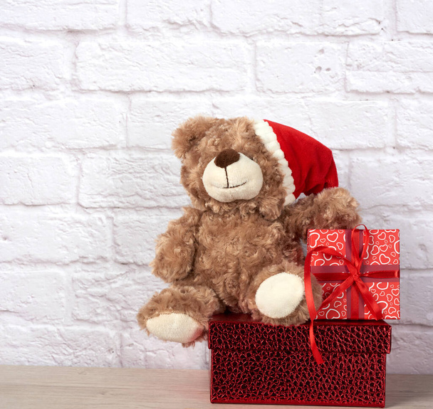 плюшевый мишка в шляпе Санта-Клауса и коробке, завязанной красной лентой, на рождественском фоне - Фото, изображение