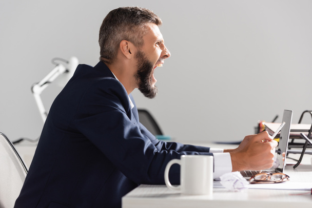 Zijaanzicht van gestresste zakenman schreeuwen terwijl het houden van potlood in de buurt van laptop en bril op wazig voorgrond in het kantoor  - Foto, afbeelding