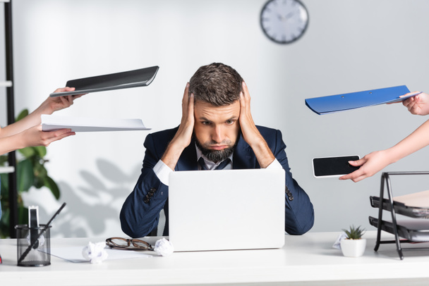 Exhausted businessman touching head near colleagues with paper folders and smartphone in office  - Photo, Image