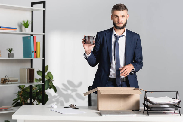 Fired businessman holding stationery near carton box, laptop and papers on table  - Photo, Image