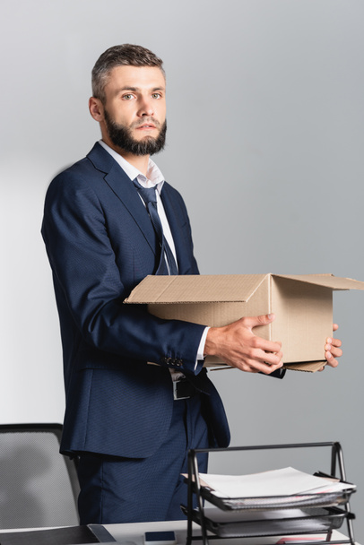 Empresario despedido con caja de cartón mirando a la cámara cerca de teléfono inteligente y papeles en la mesa  - Foto, imagen
