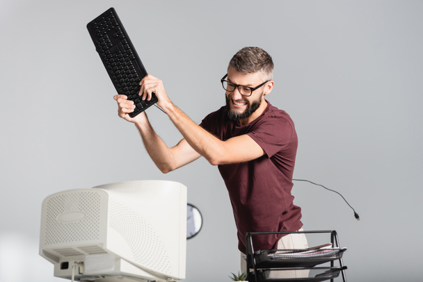 Νευρικός επιχειρηματίας κρατώντας πληκτρολόγιο υπολογιστή κοντά οθόνη σε θολή πρώτο πλάνο στο γραφείο  - Φωτογραφία, εικόνα