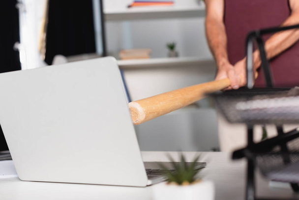 Gewassen weergave van zakenman met honkbalknuppel in de buurt van laptop met wazig voorgrond in office tafel  - Foto, afbeelding