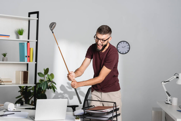Šílený obchodník drží golfovou hůl poblíž notebooku a papírnictví v kanceláři  - Fotografie, Obrázek