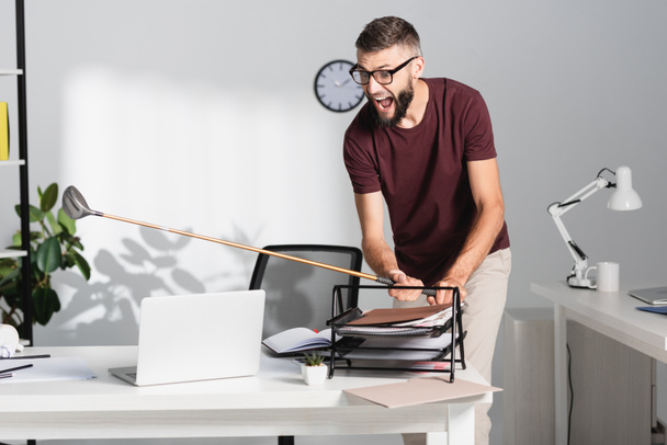 Naštvaný a křičící podnikatel drží golfovou hůl v blízkosti dokumentů a notebook v kanceláři  - Fotografie, Obrázek