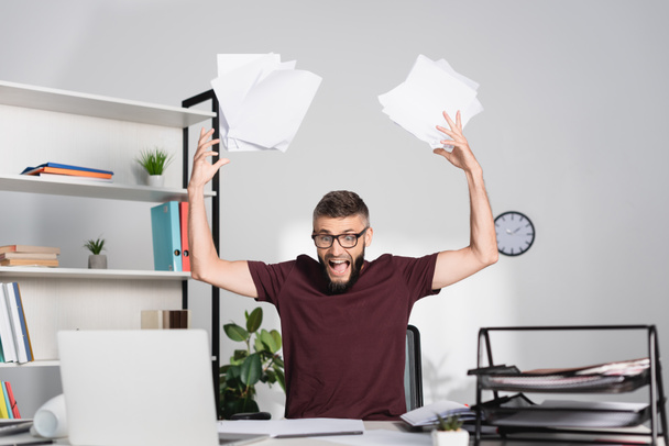 Screaming businessman throwing papers near laptop on blurred foreground in office  - Foto, Bild