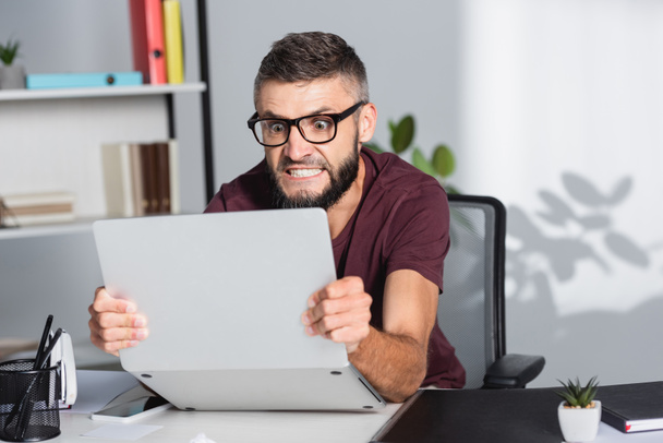 Божевільний бізнесмен тримає ноутбук біля паперу на розмитому передньому плані в офісі
  - Фото, зображення