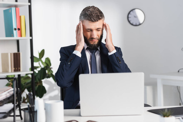 Businessman suffering from headache near laptop on blurred foreground in office  - Photo, Image
