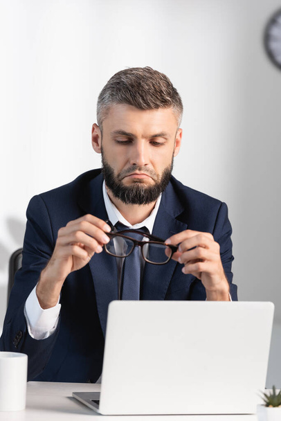 Sad businessman holding eyeglasses near laptop in office  - Fotoğraf, Görsel