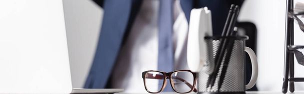Eyeglasses near stationery and laptop on office table with businessman on blurred background, banner - Foto, afbeelding