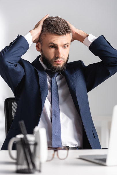 Tired businessman with hands near head looking at laptop on blurred foreground in office  - Foto, imagen