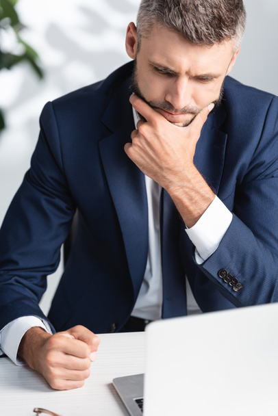 Pensive businessman with hand near chin sitting near laptop on blurred foreground  - Foto, Bild