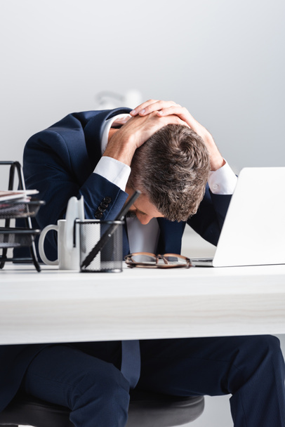 Exhausted businessman touching head near laptop and stationery on blurred foreground on table  - Foto, afbeelding