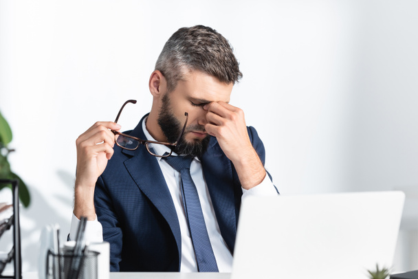 Overworked businessman holding eyeglasses and touching eyes near laptop on blurred foreground  - Photo, Image