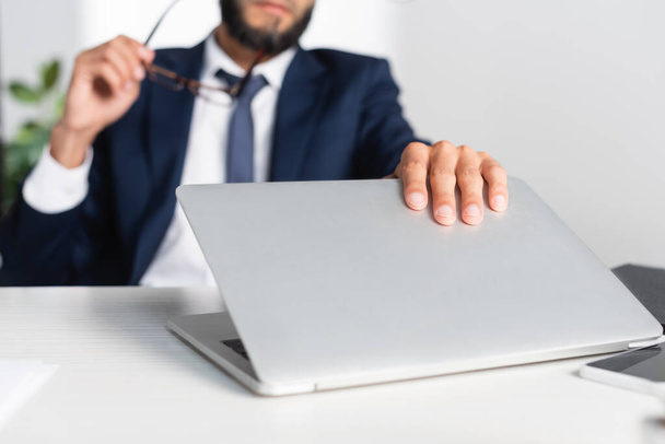 Cropped view of businessman touching laptop while holding eyeglasses on blurred background  - Zdjęcie, obraz