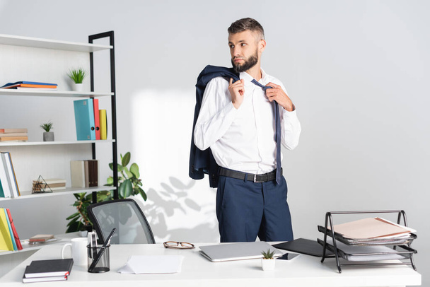 Бізнесмен торкається краватки і тримає піджак біля ноутбука і документів на столі в офісі
  - Фото, зображення