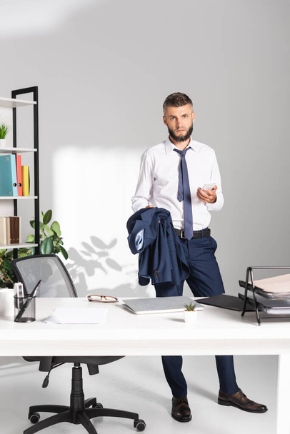 Businessman holding smartphone and jacket near papers and stationery on working table on blurred foreground  - Фото, изображение