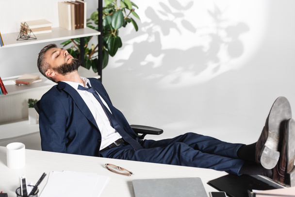 オフィスで前景にぼやけたガジェットや文房具の近くで寝ているビジネスマン  - 写真・画像