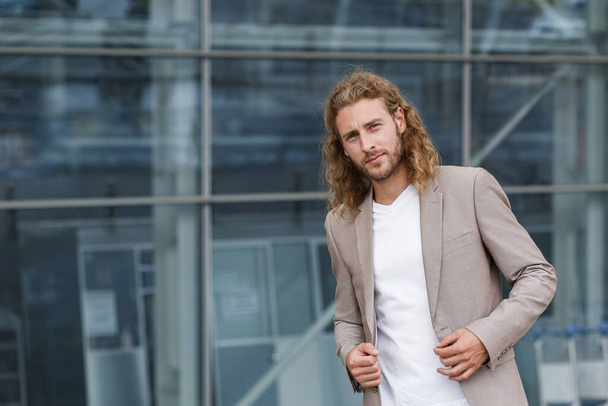 Business portrait of handsome curly smiling man with long hair in casual wear, standing on city street near business center. Confident businessman became successful - 写真・画像