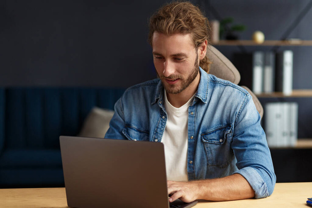 Young handsome curly smiling man with long hair working in home office with laptop. Business portrait of handsome manager sitting at workplace. Studying online, online courses. Business concept - Valokuva, kuva