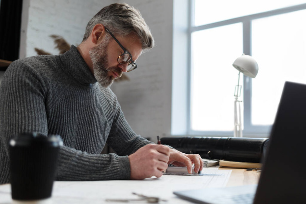Architect drawing blueprints in office. Engineer sketching a construction project. Architectural plan. Close-up portrait of handsome bearded man concentrated on work. Business construction concept - Photo, Image