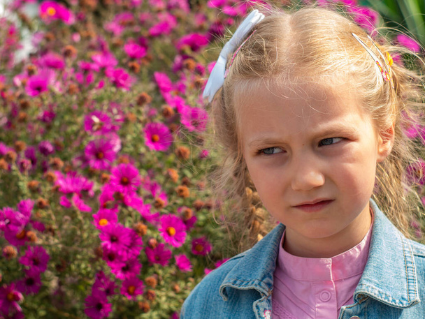 Beautiful sad blond little girl smelling flower and feeling upset. Kids pollen allergy. Sick child grimaces and sneezes on blurred nature background. Allergic rhinitis, plants blooming. - Photo, Image