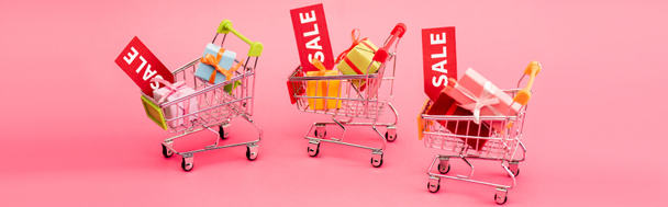 panoramic crop of colorful presents in shopping carts with sale tags on pink, black friday concept  - Photo, Image