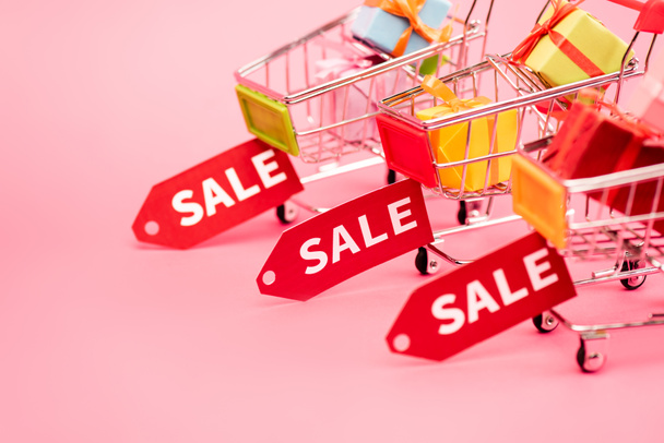 selective focus of colorful presents in shopping carts with sale tags on pink, black friday concept  - Photo, Image
