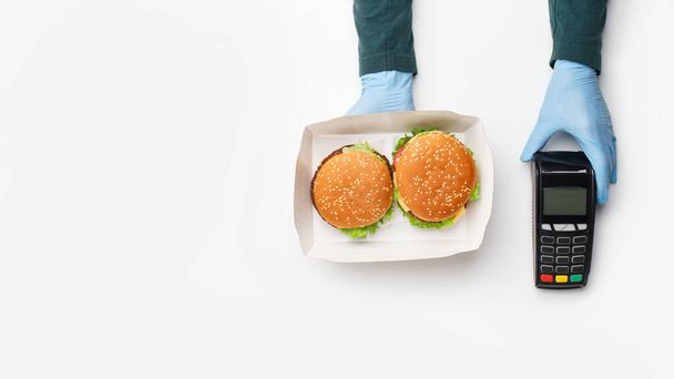 Σύγχρονες τεχνολογίες για ανέπαφες πληρωμές παραγγελιών. Σερβιτόροι χέρια σε γάντια από καουτσούκ δίνει κουτί με χάμπουργκερ και cheeseburger και τερματικό - Φωτογραφία, εικόνα