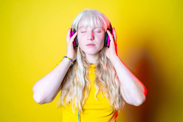 ruhige kaukasische Frau auf gelbem Hintergrund hört Musik mit Smartphone - Isoliertes junges Mädchen mit Vitiligo lächelt mit Kopfhörer - Technologie, Streaming, Musikkonzept - Foto, Bild