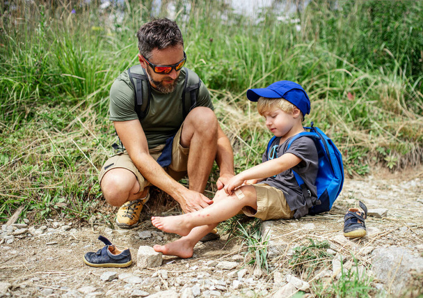 Vater mit kleinem Sohn wandert im Sommer draußen in der Natur und legt Gips aufs Knie. - Foto, Bild