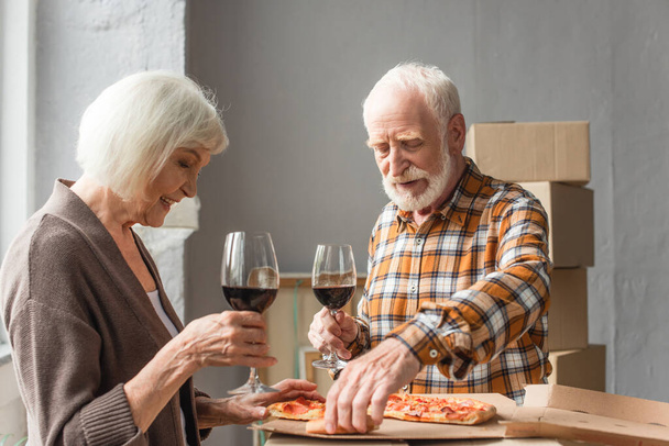 senior couple holding glasses of wine and man taking piece of pizza in new house - 写真・画像